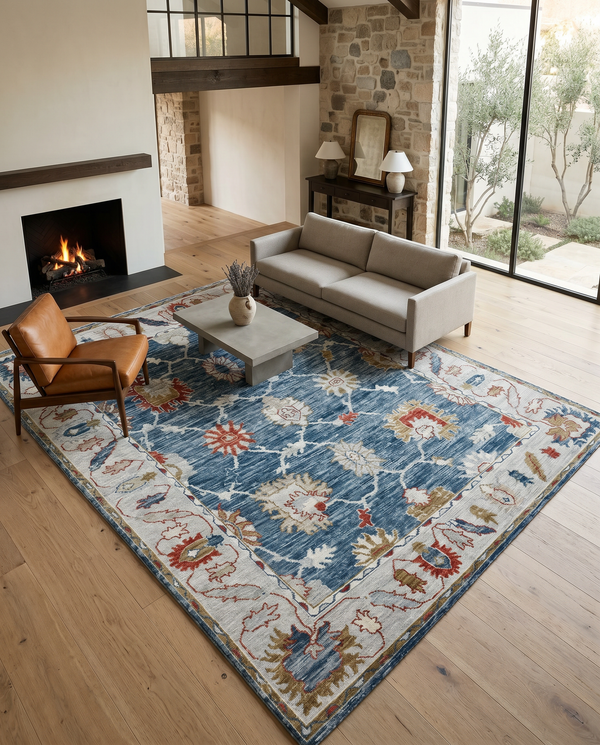 Indigo blue and ivory floral rug on pale oak flooring with a grey sofa, tan leather chair and stone fireplace.
